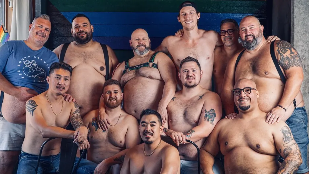 11 men smiling at the camera wihthout shirts on, standing in front of a wooden wall.